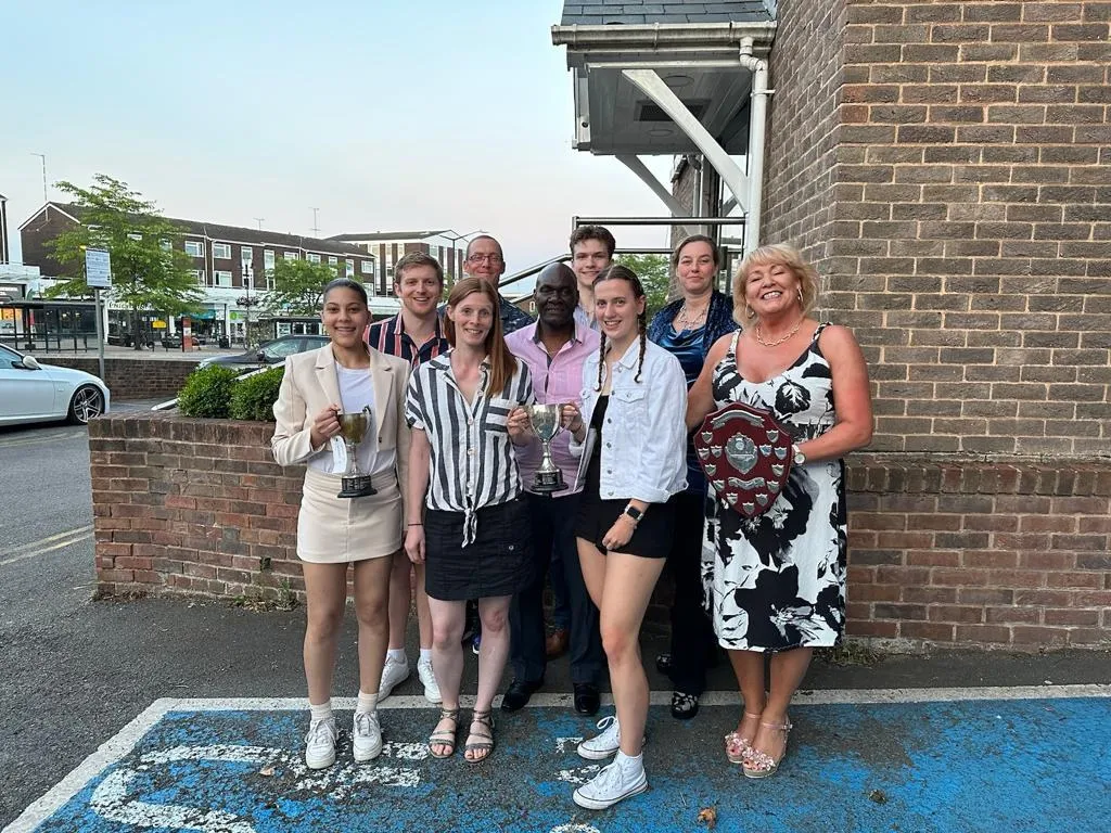 Nine club members outside at dusk, two holding silver cups and one holding an engraved shield.