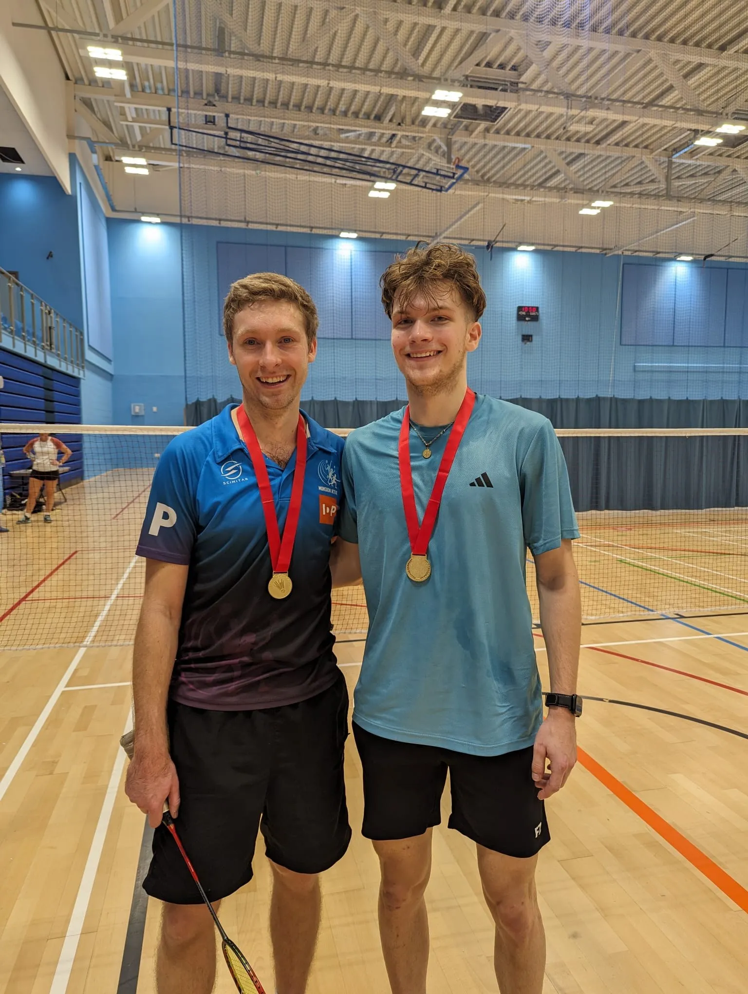 Two players in club kit on a sports hall court, both wearing gold medals on red ribbons.