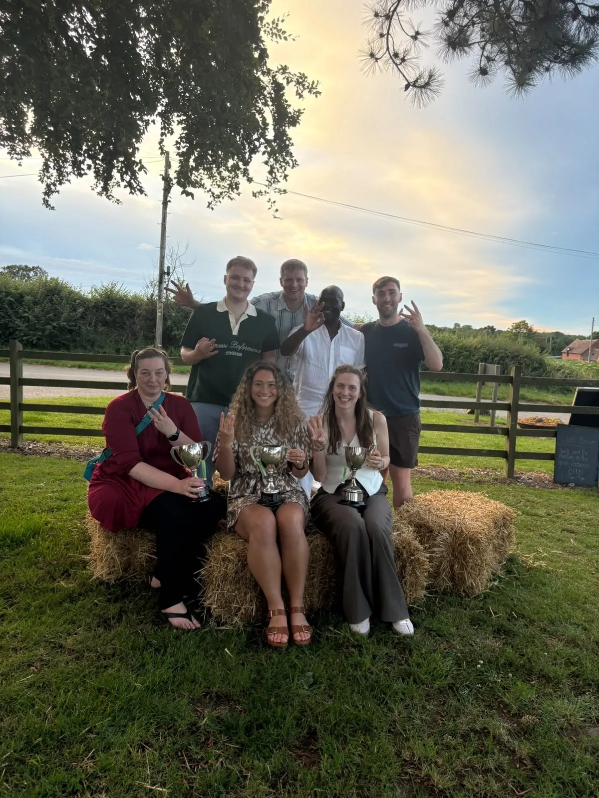 Seven club members outdoors at dusk holding three silver trophies, several of them holding up three fingers.