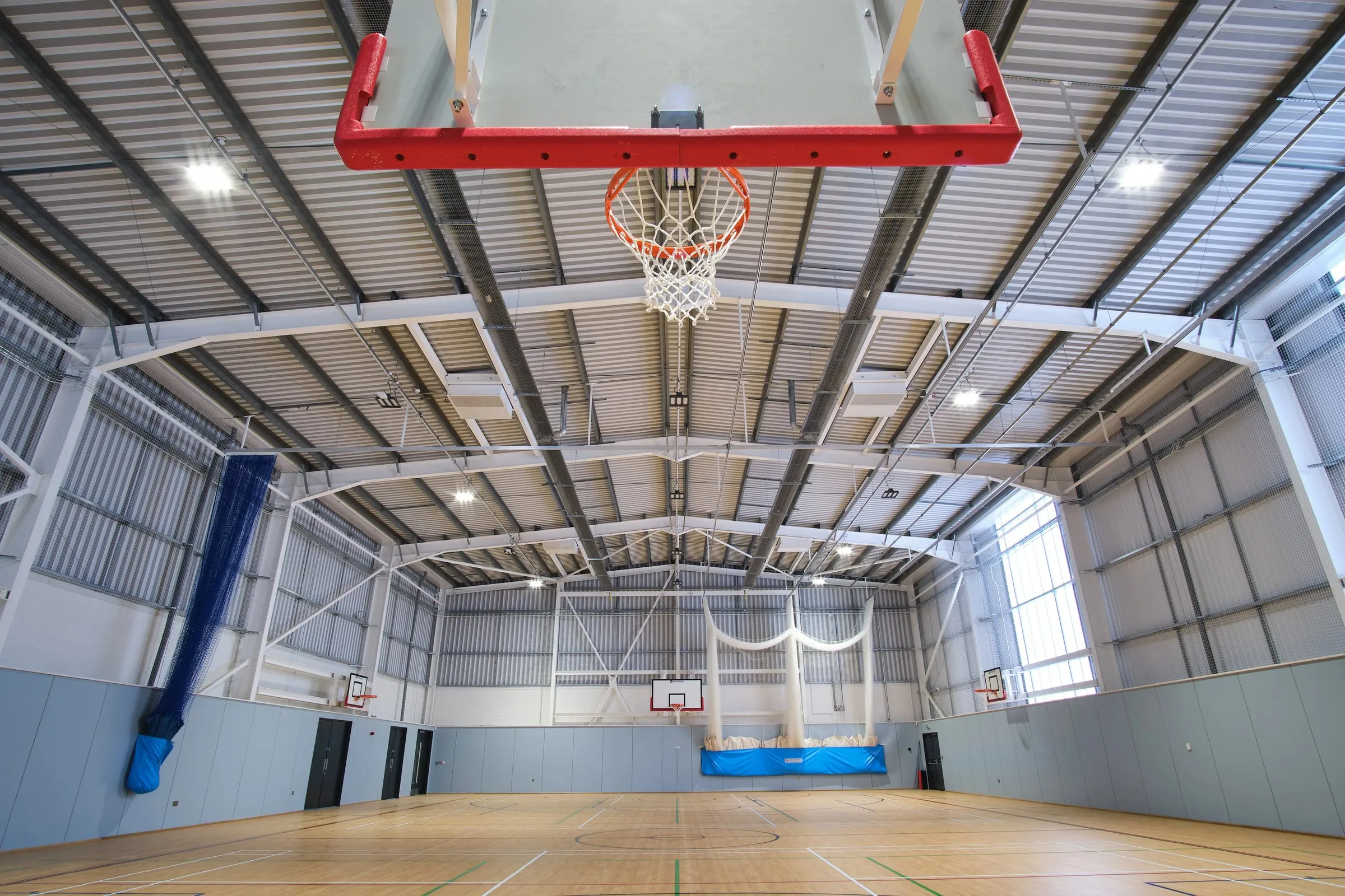 The sports hall at Bishop Ullathorne, photographed from floor level: a maple floor with court markings, a white steel roof frame and basketball backboards, with no one in the room.